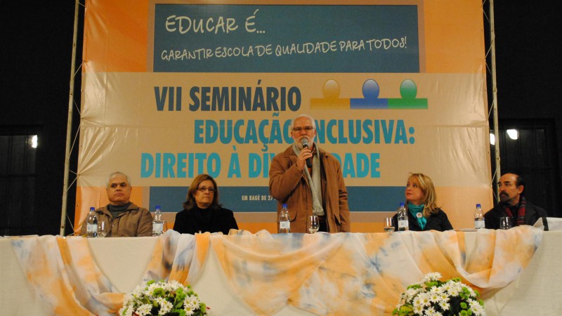 Quatro palestrantes sentados à mesa com dois arranjos de flores à frente e o banner do encontro ao fundo e no centro um palestrante de pé falando ao microfone.