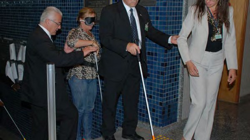 Descrição da foto da Imprensa TJRS
Cristina,Coordenadora do Programa de Acessibilidade e inclusão do TJRS, conduzindo o colega da segurança que está vendado e todos utilizando bengalas da ACERGS no topo da escada principal, identificando o piso tátil