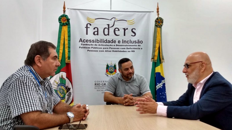 Três pessoas sentadas ao redor da mesa, estão em reunião no gabinete da presidência da FADERS. Ao fundo, da imagem, banner da FADERS, ao lado esquerdo do banner, bandeira do Rio Grande do Sul, ao lado direito bandeira do Brasil.