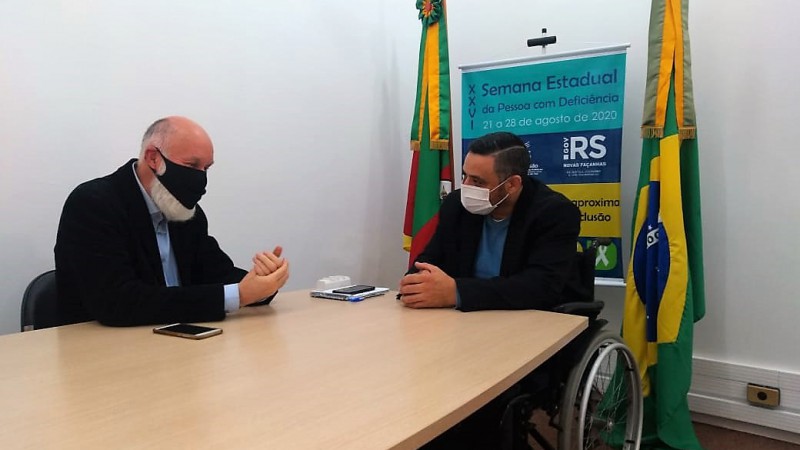 #PraTodosVerem Dois homens com os braços apoiados sobre uma mesa, um calvo e com barbas grisalhas usando máscara de proteção facial de cor preta, e outro de cabelos escuros, sentado sobre uma cadeira de rodas e com máscara de proteção facial na cor