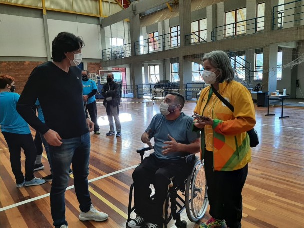 Numa quadra de esportes, três pessoas conversam. Da esquerda para a direita, o secretário de Esporte, Danrlei de Deus; o presidente da FADERS, Marquinho Lang, sentado sobre uma cadeira de rodas; e a coordenadora de Paradeporto da FADERS, Cláudia Alfama