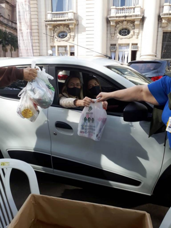 Duas mulheres dentro de um carro branco alcançam sacolas com donativos para duas pessoas das quais só aparecem os braços. Ao fundo, uma parte da fachada do Palácio Piratini.