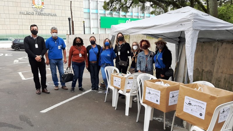 Nove pessoas, todas usando máscaras posam para foto.Parte delas está sob um gazebo. À frente, três caixas com donativos estão apoiadas sobre cadeiras de plástico. Ao fundo, o Palácio Farroupilha (Assembleia Legislativa).