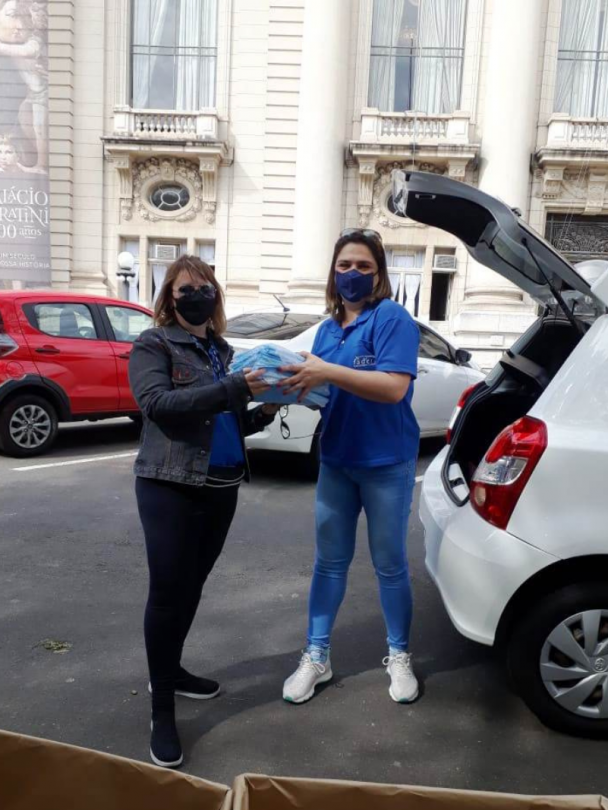 Duas mulheres em pé seguram um pacote. Ao lado, o detalhe de um carro na cor branca com o porta-malas aberto. Ao fundo, uma parte da fachada do Palácio Piratini.