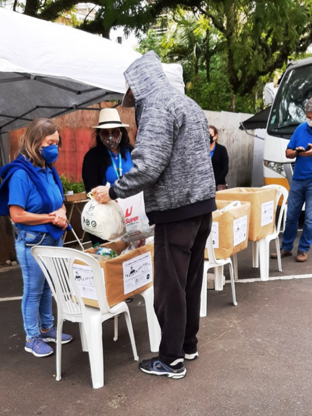 Um homem, de costas e usando capuz, alcança duas sacolas para duas mulheres. Sob as sacolas, uma caixa de papelão com donativos apoiada em cadeiras de plásticos. Ao fundo, detalhes do gazebo, do micro-ônibus e outras duas caixas apoiadas em cadeiras de plástico.