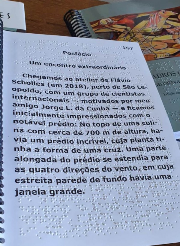 #PraTodosVerem Foto de uma página da obra em Braille. Sobre a codificação em Braille, o mesmo texto escrito em tinta preta. Segue o texto: “Posfácio. Um encontro extraordinário. Chegamos ao atelier de Flávio Scholles (em2018), perto de São Leopoldo, com um grupo de cientistas internacionais – motivados por meu amigo Jorge L. da Cunha – ficamos inicialmente impressionados com o notável prédio. No topo de uma colina com cerca de 700m de altura, havia um prédio incrível, cuja planta tinha a forma de uma cruz. Uma parte alongada do prédio se estendia para as quatro direções do vento, em cuja estreita parede de fundo havia uma janela grande. Ao fundo, a capa do volume em braille.