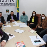 #PraTodosVerem Oito pessoas, três homens e cinco mulheres, estão sentados ao redor de uma mesa retangular. Ao fundo, o banner da FADERS entre as bandeiras do Rio Grande do Sul e do Brasil.
