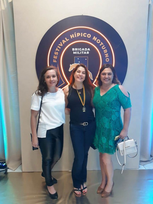 Três mulheres posam para foto em frente à logomarca do 63º Festival Hípico Noturno. Da esquerda para direita, Alane Nerbass Souza, Eva Loreni de Castilhos e Alexsandra Paz Araújo. Eva tem em seu pescoço a medalha recebida no evento.