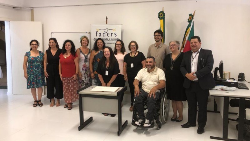 Doze pessoas, nove mulheres e três homens, posam para a foto. Todos estão em pé, exceto o presidente da FADERS, Marquinho Lang, que está sentado sobre cadeira de rodas. Ao fundo, o banner da FADERS e as bandeiras do Brasil e do Rio Grande do Sul.