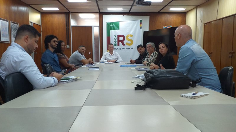 Nove pessoas estão sentadas ao redor de uma mesa retangular, conversando. Na cabeceira, o secretário Beto Fantinel, entre o presidente da FADERS, Marquinho Lang e o diretor administrativo, Romário Cruz. Ao fundo, o banner do Governo do Estado, Novas Façanhas.