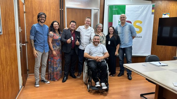 Oito pessoas posam para a foto. Todos estão em pé, com exceção do presidente da FADERS, sentado sobre cadeira de rodas. Atrás, o banner do Governo do Estado, Novas Façanhas.