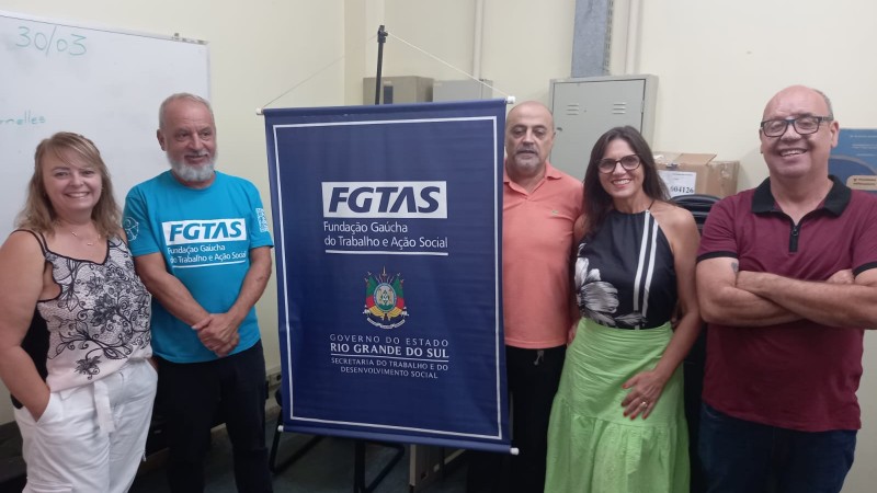 Duas mulheres e três homens posam ao redor de um banner com a logomarca da FGTAS e o brasão do Governo do Estado.