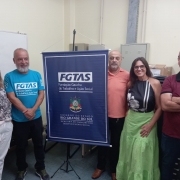 Duas mulheres e três homens posam ao redor de um banner com a logomarca da FGTAS e o brasão do Governo do Estado.