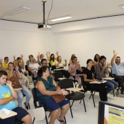 Cerca de 30 pessoas, mulheres e homens, estão sentados em cadeiras escolares. Eles acenam com ambas mãos levantadas.