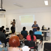 A diretora técnica da FADERS, Miriam Wornicow, está em pé, de frente para o público. À esquerda, o intérprete de Libras e uma projeção na parede com a programação da Semana de Conscientização da Síndrome de Down. Ela observada pelo palestrante, que está em pé, à direita.