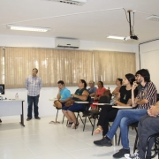 Em foto lateral, a chefe da Unidade de Eventos Técnicos, Patricia Rodrigues, está em pé, falando para o público. Também em pé, o palestrante Charlie Trellez Severo a observa. Sentado em cadeira de rodas, em primeiro plano, o presidente da FADERS, Marquinho Lang.