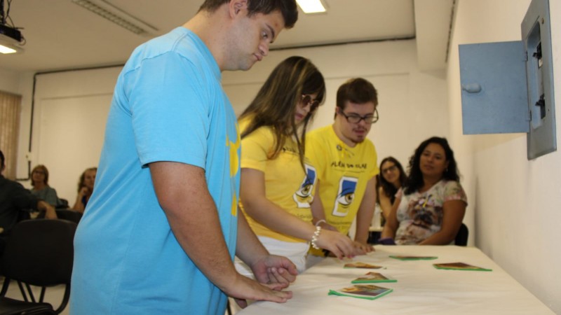 Três jovens com Síndrome de Down estão em pé, em frente a uma mesa, montando peças de um quebra-cabeças. O público, ao fundo, os observa.