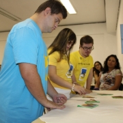 Três jovens com Síndrome de Down estão em pé, em frente a uma mesa, montando peças de um quebra-cabeças. O público, ao fundo, os observa.