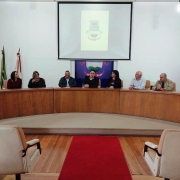 Nove pessoas, quatro mulheres e cinco homens, estão sentados atrás de uma bancada. Ao fundo, o brasão do município está projetado em uma tela. À direita, as bandeiras do Rio Grande do Sul, do Brasil e de Bagé.