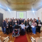 28 pessoas posam para a foto. Todos em pé, exceto o presidente da FADERS e um jovem, sentados em cadeiras de rodas. Algumas pessoas seguram certificados. Ao fundo, está projetado o brasão do município.