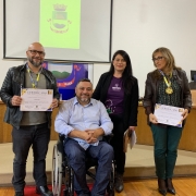 Sentado em cadeira de rodas, o presidente da FADERS posa para a foto entre um homem e duas mulheres em pé. O homem e uma das mulheres seguram certificados de participação no Praia Acessível.