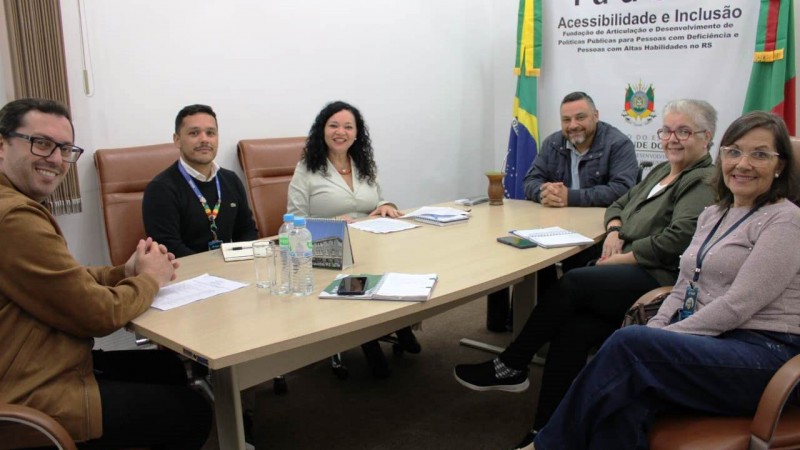 Três mulheres e três homens, sentados ao redor de uma mesa, posam para a foto. Ao fundo, o banner da FADERS entre as bandeiras do Brasil e do Rio Grande do Sul.