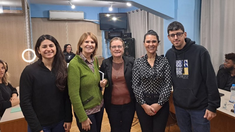 Quatro mulheres e um homem posam em pé para a foto na CIL Porto Alegre. Atrás deles, duas mulheres e um homem estão sentados atrás de mesas com computadores. As mulheres usam fones de ouvidos.