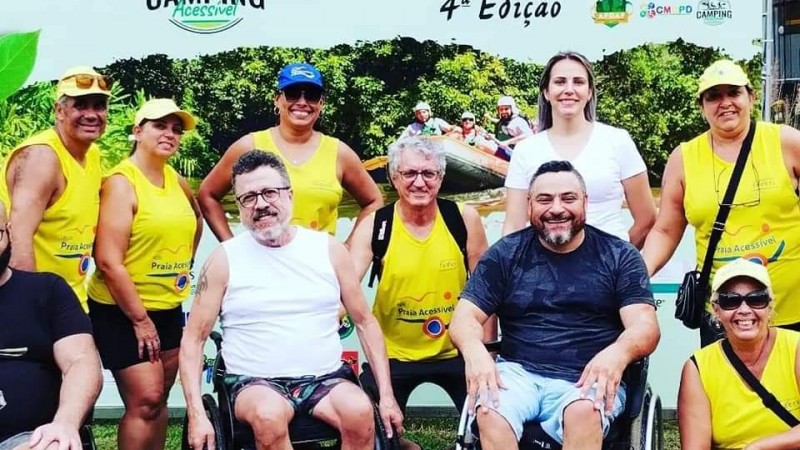 foto tirada em frente ao painel do evento Camping Acessível 2023, além da logo do evento há a frase: Bem Vindo! 4ª edição, o painel é em verde claro e as escritas em verde e reto. Em frente em pé 5 servidores da Faders com as camisetas amarelas do programa Rede Praia Acessível, á frente o presidente da Fadersde camiseta azul em uma cadeira de rodas, ao lado outro cadeirante grisalho de camiseta branca e do ouro lado mais uma funcionária da Faders abaixada com a camiseta do Rede Praia Acessível