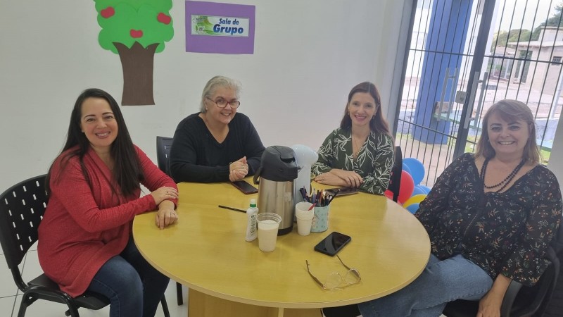 Fotoo sala com paredes em branco, ao fundo janela com cortina em azul, alguns cartazes na parede ao centro mesa redonda em madeira e ao redor cadeiras pretas, onde estão sentadas 4 mulheres, 3 servidoras da faders e a coordenadora do Cas Teacolhe de Soledade.