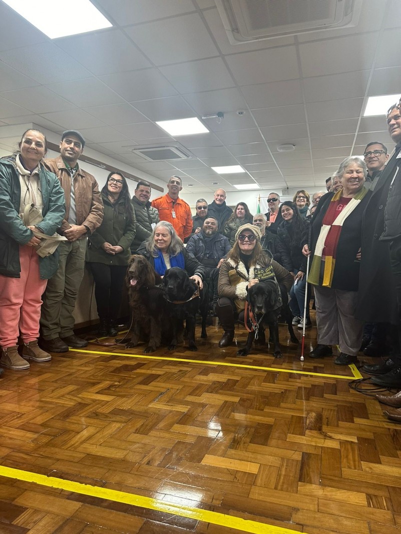 foto do evento - no centro os três cães-guia presentes: Darwuin marrom e Cobe e Estrela pretos, acompanhados de suas tutoras, as deficientes visuais Olga e Marilena, atrás delas em cadeiras de rodas o presidente da Faders e o presidente do Coeped, em pé o Major Moreno representando o Comando dos Bombeiros Militar do RS, o ex-presidente a Faders Backof, o professor Leonardo do IFF SC, diretoria técnica da Faders e demais participantes da reunião