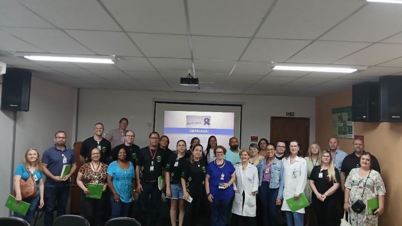 Foto do grupo que participou da palestra, ao centro a palestrante, ao fundo uma parede branca e uma tela com apresentação. Fim da descrição