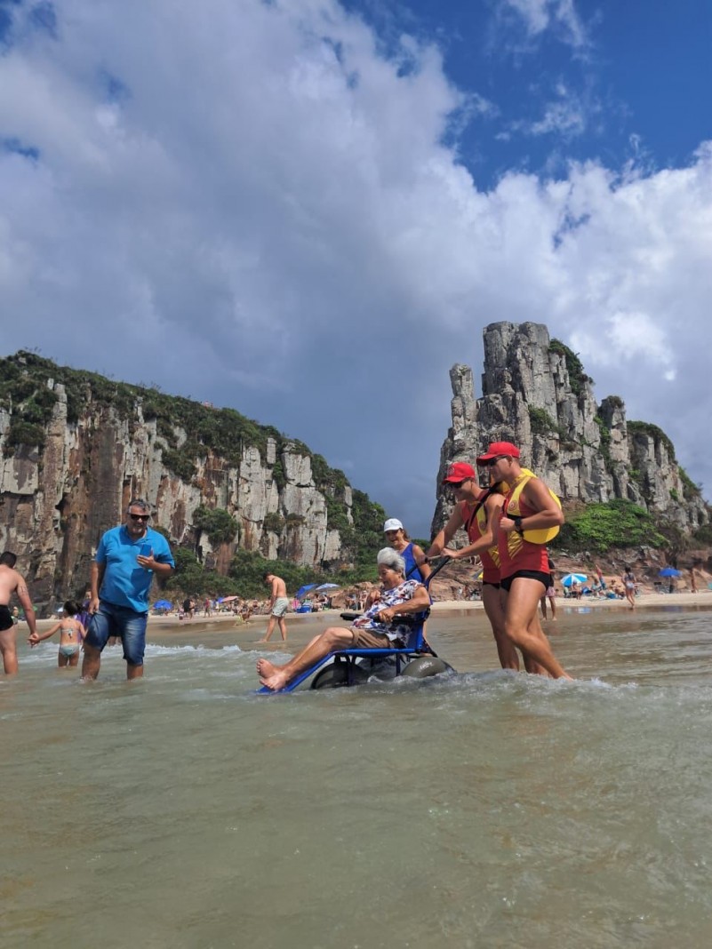 Foto- dois guarda vidas acompanham uma senhora na cadeira anfíbia em um banho de mar.