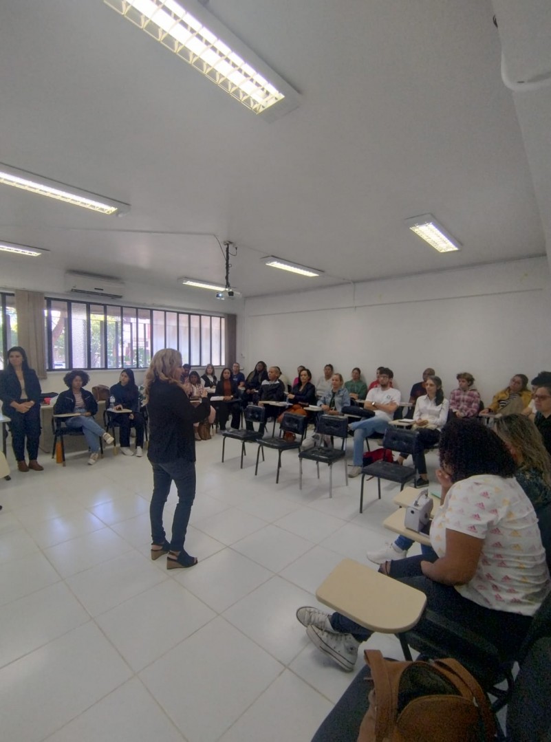 Foto - turma Morretes - uma sala com paredes e piso brancos, ao redor em formato de U estão distribuídas cadeiras de estudos com a mesa acoplada onde estão os alunos, ao centro a instrutora dando orientações. Ela é loira cabelos encaracolados até o ombro, esta na foto meio de lado e veste calça jeans um sapato e uma camisa pretos. Fim da descrição