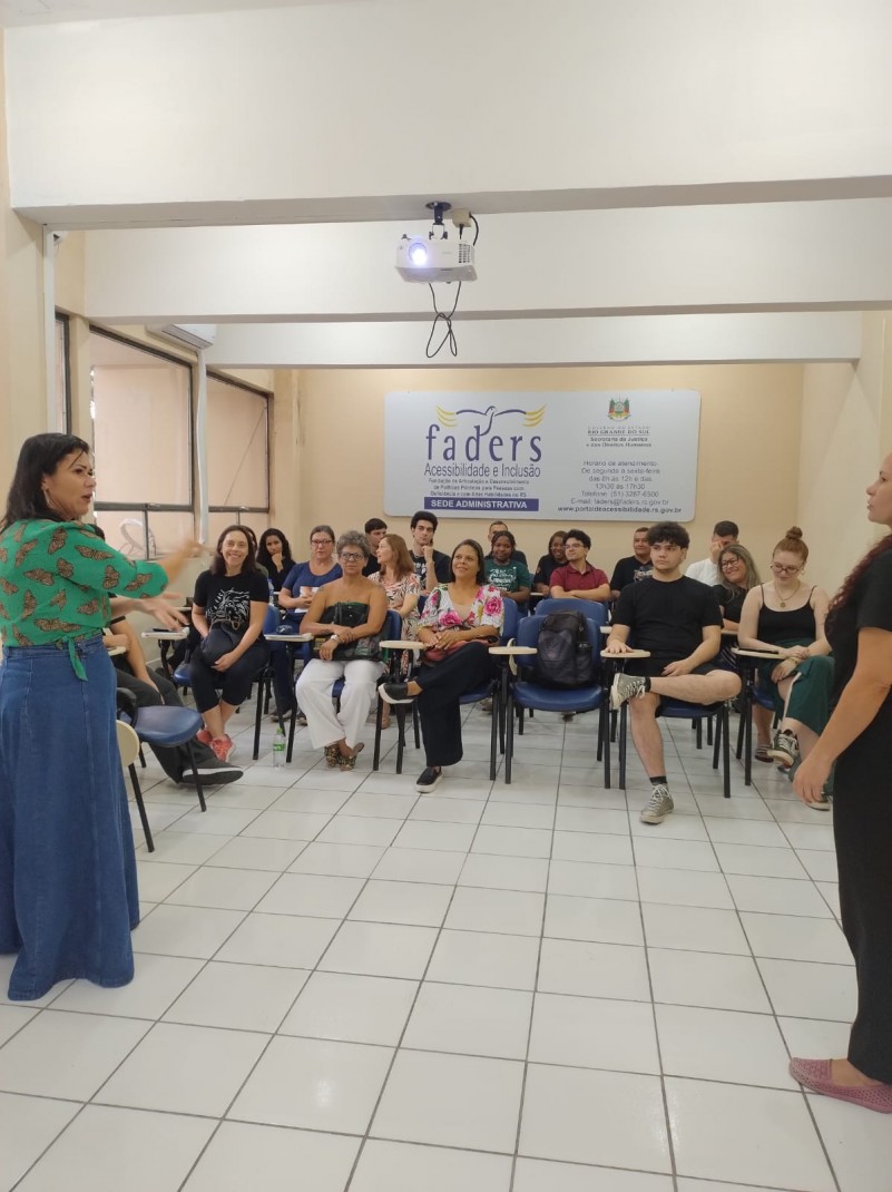 Foto- turma Sede/Centro - uma sala com piso branco e paredes em um tom de bege, ao fundo uma placa com a Logo da Faders. Os Alunos encontra-se sentados em cadeiras com centos azuis e a mesas acopladas em bege, na frente a instrutora de Libras proferindo a aula. Ela tem cabelos pretos e lisos ate o ombro, esta de lado, com uma saia longa jeans e um camisa cerde. Fim da descrição