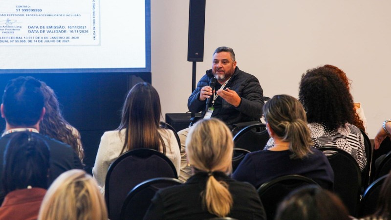 foto - publico de costas sentado em cadeiras com encostos preto, e em frente a uma tela onde está a apresentação da Ciptea, o presidente da Faders, em sua cadeira de rodas, e com o microfone em uma das mãos e a Carteira de identificação da Pessoa com Especto Autista em outra, fala sobre a importância do documento. Fim da descrição