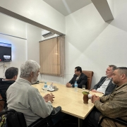 Foto- sala com paredes brancas, ao centro uma mesa com taqmpa em madeira clara, ao redor cadeiras em couro marrom, na ponta da mesa em sua cadeira de rodas o presidente da Faders, a dua esquerda o presidente do Coepede também em sua cadeira de Rodas, ao redor deles os demais três participantes da reunião. Em frente a mesa uma televisao transmite a participação por videoconferência de um delegado do Coepede. Fim da descrição