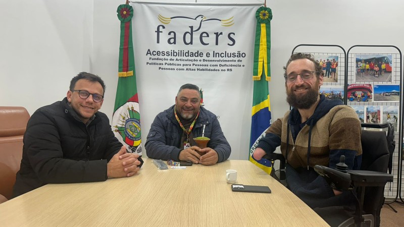 foto- ao fundo parede branca, com o banner da Faders e as bandeiras do RS e Brasil, mais a direita da pra ver um painel de fotografias com a sprincipais atividades da Fundação. Em primeiro plano ao redor de uma mesa com tampa de madeira clara esta na ponta o presidente da Faders em sua cadeira de Rodas, do lado esquerod o diretor geral em uma cadeira com encosto marrom em couro e do direito o diretor de inclusão também em sua cadeira de rosas da Secretaria de Justiça, Cidadania e Direitos Humanos do Rio Grande do Sul. Fim da Descrição