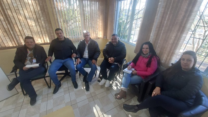 Foto- em uma sala com piso de cores claras e janelas ao fundo da parede, em primeiro plano estão 6 pessoas sentadas em cadeiras com encosto preto, ao centro o presidente da Faders em sua cadeira de rodas. Fim da descrição
