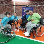 FOTO- em uma quadra de esportes estão o presidente da Faders e mais 3 meninos com idades variadas em suas cadeiras de rodas em um circulo e ao centro eles seguram uma bola de basquete. Os meninos estão de camiseta azul e abrigo cinza e o presidente da Faders de camiseta verde e abrigo cinza. Todos sorriem com expressão de felicidade. Fim da descrição