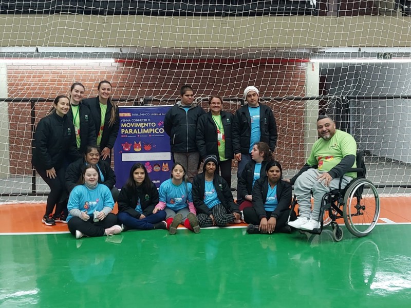 FOTO - Presidente da Faders posa para foto com crianças e adultos que participaram do Festival do Paradesporto em Erechim. Estão em uma quadra esportiva e ao fundo preso da Rede um banner do Movimento Paralímpico. Fim da descrição