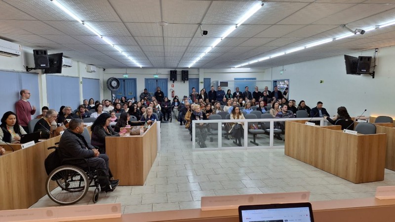 Foto- auditório da câmara de Vereadores de Santo Antônio da Patrulha, foto tirada da mesa da presidencia, que pega de frente as mesas dos vereadores onde está a equipe da Fader e o presidente, em frente as cadeiras azuis dos visitantes onde estão os participantes do evento sentados, e alguns em pé no fundo. A sala está lotada. Fim da descrição