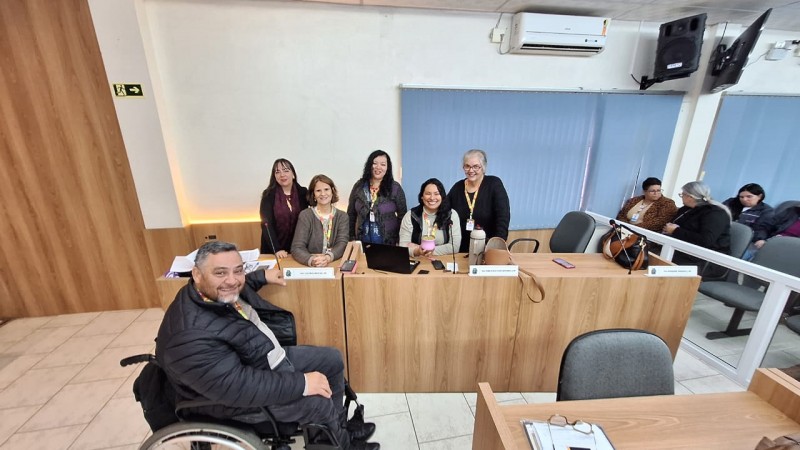 Foto- no auditório da Câmara de Vereadores de Santo Antônio da Patrulha, em uma mesa em madeira, estão a diretora técnica da faders e as coordenadoras de Pesquisa, Acessibilidade, Políticas Públicas e Capacitação da Faders, em frente a mesa na sua cadeira de rodas o presidente da Faders, O Piso é bege claro do local e as paredes brancas com cortinas azuis. Fim da descrição.