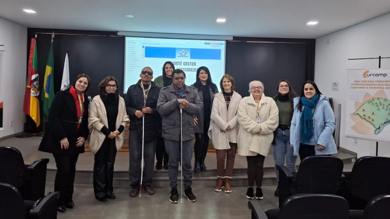 Foto - sala com paredes brancas, ao fundo parede preta com um telão no centro onde esta sendo projetado o material debatido na  Oficina. Na frente em primeiro plano e todos em pé, a equipe que faz parte do Comitê Gestor Municipal do Sele de Acessibilidade em Bagé, a diretora técnica  e a coordenadora de Acessibilidade, ambas da  Faders. Fim da descrição