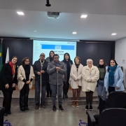 Foto - sala com paredes brancas, ao fundo parede preta com um telão no centro onde esta sendo projetado o material debatido na  Oficina. Na frente em primeiro plano e todos em pé, a equipe que faz parte do Comitê Gestor Municipal do Sele de Acessibilidade em Bagé, a diretora técnica  e a coordenadora de Acessibilidade, ambas da  Faders. Fim da descrição