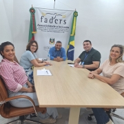 A imagem mostra cinco pessoas sentadas ao redor de uma mesa retangular de madeira, em uma sala de reuniões iluminada. As duas à esquerda ( a coordenadora de Acessibilidade da Faders e uma servifora de Capão da Canoa) sentadas em cadeiras de couro marrom. Ao fundo, há um banner da Faders – Acessibilidade e Inclusão, acompanhado das bandeiras do Brasil e do Rio Grande do Sul. Todos estão posicionados de forma frontal ou lateral para a câmera, sorrindo, em um ambiente formal e acolhedor, com documentos e uma garrafa de água sobre a mesa.No centro o presidente da Faders em sua cadeira de Rodas, e ao lado dele o servidor de capão de canoa tbm em cadeira de rodas, ao lados des a direita a outra servidora de Capão da Canoa em uma cadeira em couro marrom.
