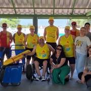 A imagem mostra um grupo de cerca de 18 pessoas reunidas sob uma estrutura coberta, em um espaço externo próximo a uma piscina. Na parte frontal, há duas cadeiras anfíbias de cor azul e um flutuador amarelo utilizado por guarda-vidas. O grupo é composto por homens e mulheres, alguns usando camisetas e bonés amarelos com a marca do programa Operação Verão Total, os guarda-vidas de uniforme vermelho e amarelo, enquanto outros vestem roupas casuais. Algumas pessoas estão em pé e outras agachadas ou sentadas,e o presidente da Faders ao centro em sua cadeira de rodas,  formando uma pose coletiva para a foto. O ambiente ao fundo mostra área de lazer aquática com piscinas e estruturas de parque. 