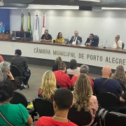 Foto feita do fundo do auditório da Camara de Vereadores de Porto Alegre, a frente em cadeiras pretas várias pessoas acompanham a abertura do Seminário. Em frente ao publico na mesa oficial as autoridades que participaram da abertura oficial do evento, no canto esquerdo de camieta preta com a logo da Faders o presidente da Funação. O chão é revestido de carpete marrom escuro e as paredes são brancas.