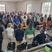 Fotografia em ambiente interno, no audit&oacute;rio da C&acirc;mara de Vereadores de Santiago, com grande n&uacute;mero de pessoas reunidas. A imagem mostra dezenas de participantes em p&eacute;, organizados entre fileiras de cadeiras, voltados para a parte frontal da sala.
No primeiro plano, aparecem algumas pessoas de costas, possivelmente autoridades ou organizadores, posicionadas &agrave; frente do p&uacute;blico. Entre elas, h&aacute; um homem utilizando cadeira de rodas. Tamb&eacute;m &eacute; poss&iacute;vel ver um projetor sobre uma mesa, indicando que o espa&ccedil;o &eacute; utilizado para apresenta&ccedil;&otilde;es.
O p&uacute;blico &eacute; diverso, composto por homens e mulheres de diferentes idades, vestindo roupas casuais e formais. O ambiente possui paredes claras, janelas grandes nas laterais que permitem entrada de luz natural, e cadeiras dispostas em fileiras.
A cena transmite um momento coletivo de aten&ccedil;&atilde;o e participa&ccedil;&atilde;o, durante a abertura do F&oacute;rum da Faders.