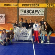 Foto feita no Gin&aacute;sio onde foi realizado o Desafie-se, a frente a servidora Claudia segura medalhas e trofeus do evento, ao lado dela uma das participantes acocoradas, ao lado todos os demais participantes em p&eacute;  com a bandeira da Funda&ccedil;ao. 