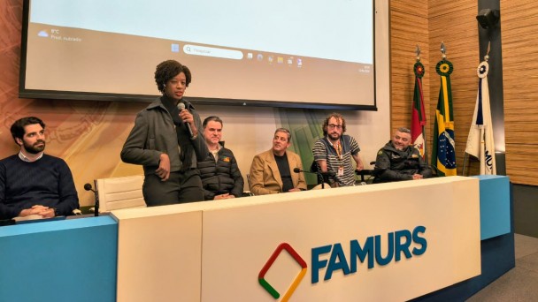 foto feita no audit&oacute;rio da Famurs, na frente a mesa princijpal, no palco com a logomarca grande da FAMURS, sentadas em cadeiras brancas oiu em cadeiras de rodas  as autoridades que participaram da abertura do evento, em p&eacute; com microfone em maos falando esta a secret&aacute;ria nacional dos Direitos da Pessoa com Defici&ecirc;ncia, Isadora Nascimento